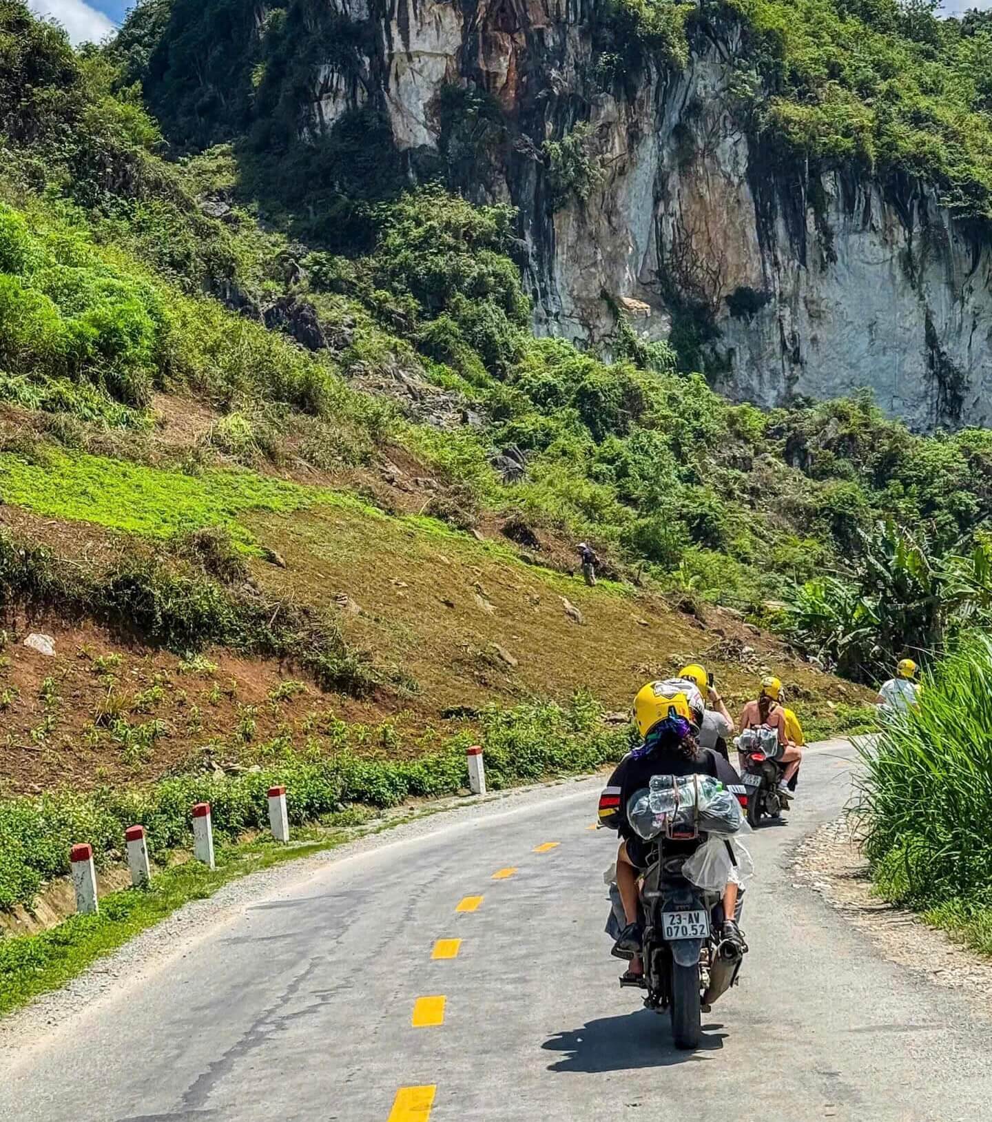 A group of travelers and their local Easy Rider guides wearing matching yellow helmets, safely riding motorbikes along a sunny, winding mountain road on the Ha Giang loop, with luggage securely strapped to the back of the bikes.